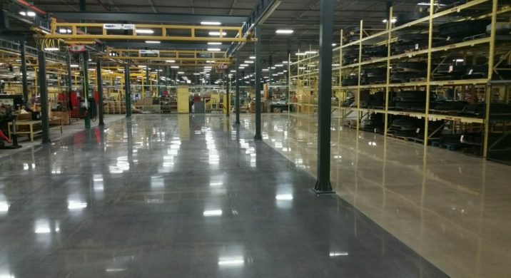 High-gloss polished concrete floor in a large industrial manufacturing facility with steel columns, overhead cranes, and storage racks, showcasing a finished commercial concrete polishing project designed for durability and heavy equipment traffic.